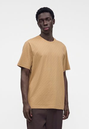 Ung mand står iført en tekstureret beige kortærmet t-shirt og mørkebrune bukser mod en ensfarvet lys baggrund.