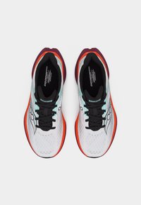 Ζευγάρι λευκά αθλητικά παπούτσια με μαύρη κορδόνια, πορτοκαλί και μωβ σόλες και τιρκουάζ λεπτομέρειες, από πάνω σε λευκό φόντο.