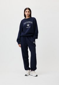 Sweatshirt bleu marine et ensemble de jogger assorti en tissu doux, avec un logo graphique en blanc. Les poignets et les chevilles sont côtelés.