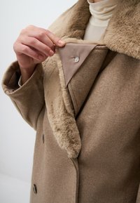Cappotto di lana beige con un colletto in pelliccia sintetica. L'interno del colletto presenta un pannello di tessuto liscio e un dettaglio con bottone. Frontale a doppi petti con bottoni.