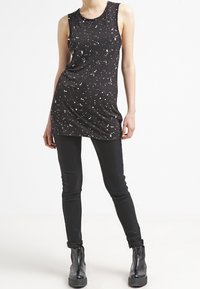 Svart ärmlös topp med ett prickigt vitt mönster, kombinerad med smala svarta byxor och ankelstövlar, mot en ren vit bakgrund.