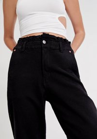 Svarta denimbyxor med elastisk midja och silverfärgad knappstängning. Har sidofickor och kopparfärgade detaljer. Slät textur.