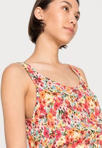 Top sem mangas com padrão floral, exibindo uma mistura de flores vermelhas, amarelas e verdes sobre um fundo claro. Tecido suave com um corte ligeiramente solto.