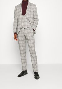 Kolme osaa sisältävä ruutukuosi puku harmaana ja valkoisena, burgundinpunainen poolopaita alla. Kiiltävät mustat loafersit viimeistelevät asun.