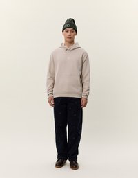 Beige hettegenser med frontlomme, kombinert med marineblå kordfløyelsbukser med et subtilt mønster. Modellen har på seg en mørkegrønn lue.