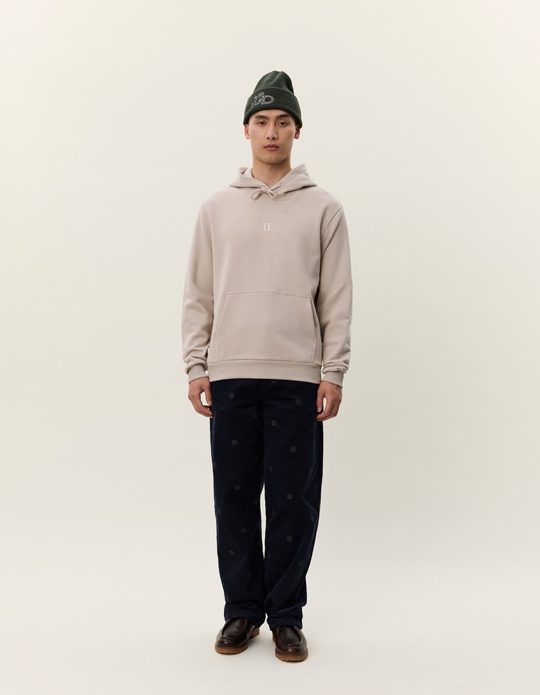 Beige hettegenser med frontlomme, kombinert med marineblå kordfløyelsbukser med et subtilt mønster. Modellen har på seg en mørkegrønn lue.
