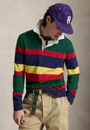 Άτομο που φοράει πολύχρωμη ριγέ ράγκμπι μπλούζα, μπεζ παντελόνι με ριγέ ζώνη και μωβ καπέλο με το γράμμα "R" κοιτάζοντας προς τα κάτω.