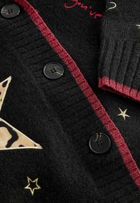 Cárdigan negro de punto con un ribete texturizado rojo, que presenta una gran estrella color marrón claro con estampado de leopardo, acentos de estrellas doradas y botones negros.