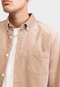 Beige Cord-Shirt mit Button-Down-Kragen, vertikal gerippter Textur, Fronttasche und weißen Knöpfen. Über einem weißen T-Shirt getragen.