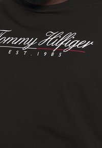 Černé bavlněné tričko s bílým kurzívním logem "Tommy Hilfiger" a nápisem "EST. 1985" v malých písmenech pod ním, s červenou akcentní linkou.