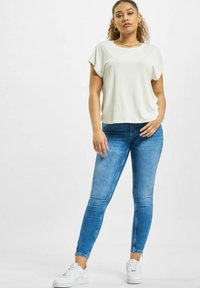 Kremfarget kortermet topp kombinert med tettsittende blå jeans og hvite sneakers. Jeansene har en teksturert finish og glidelåsdetaljer.