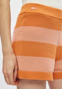 Main reposée à côté de shorts en tricot avec de larges rayures horizontales orange et rose clair et un petit logo de crocodile sur la taille.