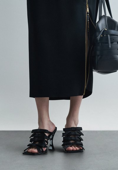 Des pieds portant des sandales à talons hauts noires avec plusieurs brides en forme de nœud, se tenant sur un sol gris, associés à une jupe noire et un sac à main noir avec des détails dorés.