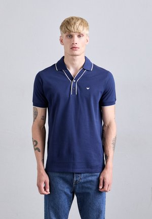 Marineblaue Polohemden aus Baumwolle mit geripptem Kragen, weißen Paspeln und Logo-Stickerei; getragen mit hellblauen Jeans, kurze Ärmel.