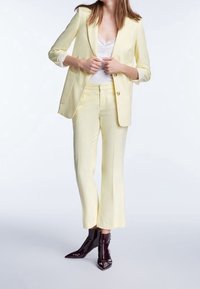 Keltainen bleiseri, jossa on läpät, ja yhteensopivat räätälöidyt housut, molemmat silkkisessä viimeistelyssä. Pukeutunut valkoisen pitsitopin päälle ja yhdistetty kiiltäviin mustiin nilkkabootseihin.