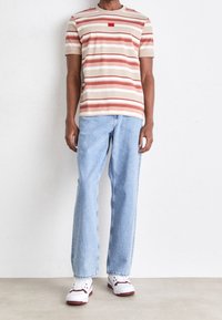 Stribet t-shirt i beige og rød, parret med lyseblå jeans. Sneakers er hvide med røde accenter. Afslappet, løs pasform.