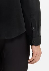 Μαύρο πουκάμισο με μακριά μανίκια και υφή στο ύφασμα, με κουμπωτά μανσέτες και ίσια τελειώματα. Το πουκάμισο συνδυάζεται με σκουρόχρωμο παντελόνι.