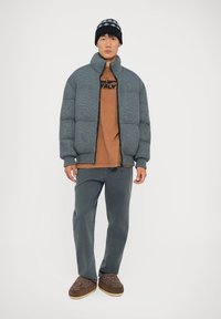 Grå quiltet pufferjakke, brun t-shirt med print, grå bukser og brune slippers; model med en mønstret hue.