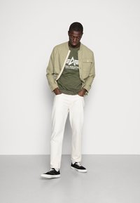 Olivgrünes T-Shirt mit Logo unter einer hellen khakifarbenen Jacke, weißen Denim-Hosen und schwarzen Sneakers mit weißen Sohlen, vor einem schlichten Hintergrund.