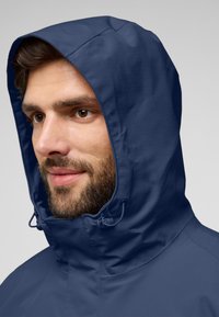 Giacca a cappuccio blu navy con un tessuto liscio e testurizzato. Corda regolabile al collo. Primo piano di una persona che indossa la giacca.