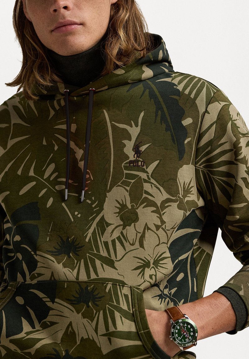 Kapuzensweatshirt in olivgrün mit floralem und Blattmuster. Verfügt über eine Kängurutasche und braune Kordeln, ergänzt durch eine Armbanduhr.