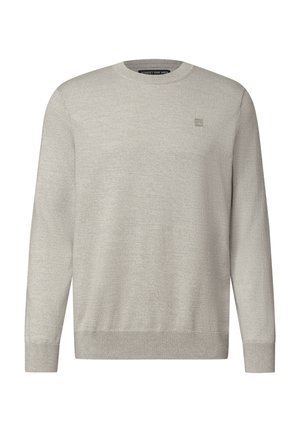Homme aux cheveux foncés et à la barbe, portant un pull gris clair à manches longues, regardant sur le côté devant un fond clair uni.