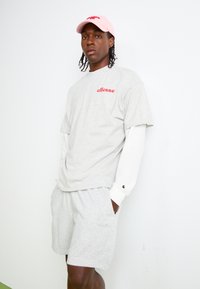 Sivý krátky rukáv tričko s červeným logom "ellesse", vrstvené cez dlhú bielu košeľu. Svetlosivé šortky, zodpovedajúci casual štýl, s vreckami. Rúžová čiapka.