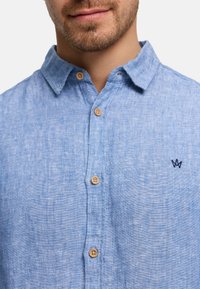 Mies, jolla on vaaleansininen napillinen paita, jossa on puiset napit ja pieni tumma kirjailtu logo rinnassa.