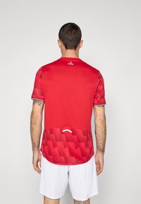 Rød atletisk trøje med korte ærmer, der har et struktureret geometrisk mønster i siderne og en rund nederste kant. Hvide detaljer og logo nær kraven.