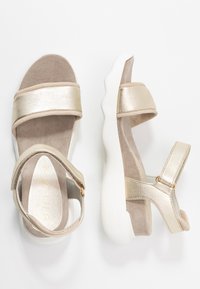 Zlaté metalické sandály se dvěma širokými popruhy, měkkou béžovou stélkou a bílou texturovanou podešví. Nastavitelný řemínek kolem kotníku se zlatým kováním.