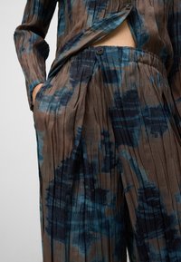 Geplooide, lange mouwen blouse en bijpassende broek van gestructureerde stof. Met een blauw en bruin abstract patroon, een zij-knopen sluiting en zakken.