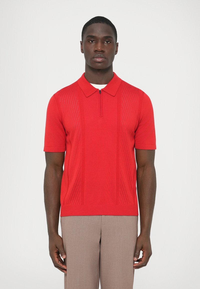 Punainen lyhythihainen poolopaita, jossa on vetoketjullinen kaulus ja teksturoidut pystysuorat raidat. Materiaali on neulosta, jossa on sileä viimeistely. Yhdistetty beigenvärisiin housuihin.