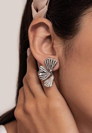Silver wavy double-leaf shaped stud earring on left ear with hand gently touching earlobe, dark hair held by beige headband.