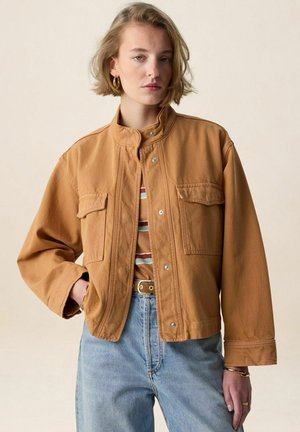 Jong persoon met kort blond haar, een bruine jas met knopen, een gestreept shirt, lichtblauwe spijkerbroek en gouden oorringen, staand in een neutrale houding.