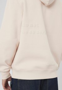 Hoddie rose clair avec un design texturé au dos, comportant les phrases « ne pas » et « ne soyez pas si sérieux » en lettres en relief. Tissu doux, coupe décontractée.