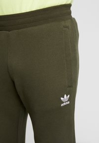 Olivgröna sweatpants i mjukt material, med dragsko i midjan och en vit Adidas trefoil-logo på vänster lår.