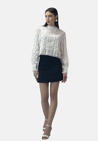 Top bianco in pizzo a maniche lunghe con intricati motivi floreali, abbinato a una gonna mini nera aderente e tacchi bianchi con cinturino alla caviglia.