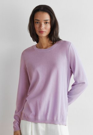 Kvinde med skulderlangt mørkt hår iført en lys lilla langærmet trøje og hvide bukser, stående foran en ensfarvet hvid baggrund.