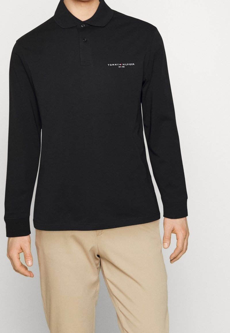 Svart långärmad polo skjorta av bomull med krage, knäppning fram och vit logotyp på vänster bröst. Slät textur, avslappnad passform.