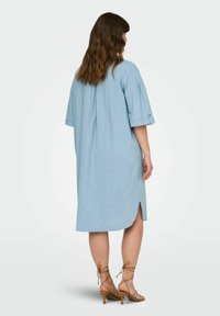 Robe en lin bleu clair à coupe ample, avec des manches courtes retroussées, des fentes sur les côtés et un pli dans le dos, associée à des talons à lanières beiges.