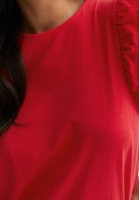 Haut rouge à manches courtes avec une texture lisse et des détails froncés aux épaules, doté d'un col rond et d'une silhouette ajustée.