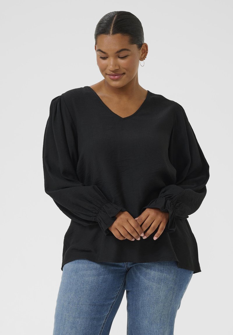 Schwarze Bluse mit V-Ausschnitt, langen Puffärmeln und gerafften Manschetten. Hergestellt aus einem leichten Stoff mit strukturierter Oberfläche. Kombiniert mit blauen Jeans.