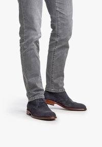 Botas de ante oscuras de hombre en color azul marino, con un diseño texturizado, punta redonda y suela de madera marrón, combinadas con jeans ajustados grises.
