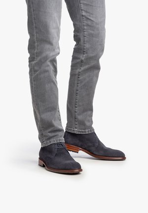 Botas de ante oscuras de hombre en color azul marino, con un diseño texturizado, punta redonda y suela de madera marrón, combinadas con jeans ajustados grises.