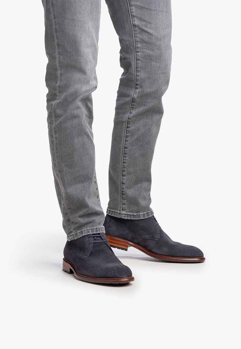 Botas de ante oscuras de hombre en color azul marino, con un diseño texturizado, punta redonda y suela de madera marrón, combinadas con jeans ajustados grises.