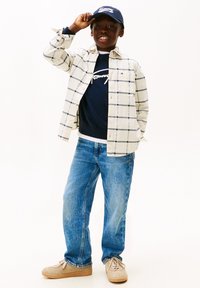 Et barn har en cremefarvet og marineblå ternet skjorte på, en marineblå sweater med logo, blå jeans og beige sneakers, mens det holder en marineblå kasket.