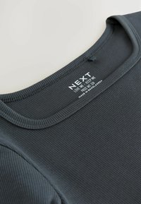 Débardeur côtelé gris foncé avec un col rond. Il présente une étiquette avec la marque "NEXT" et des instructions de lavage. Texture lisse.