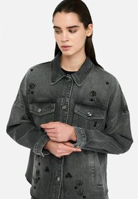 Graue Denimjacke mit lockerer Passform, doppelten Brusttaschen und floralen Perlenverzierungen. Strukturiertes Material mit Akzenten aus silberfarbigem Metall.