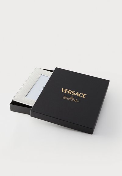 Svart ask med guld "Versace"-logotyp, innehållande en silverfärgad fotoram med en texturerad kant och ett vitt inre utrymme för ett foto.