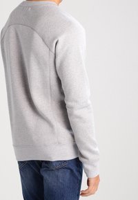 Uomo che indossa un felpa grigia chiara e jeans blu, visto di spalle su uno sfondo chiaro e semplice.